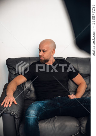 Man sitting on a gray couch in a simple indoor setting during daytime 138877253
