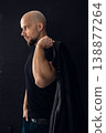 Man stands against dark background holding jacket over shoulder in casual pose 138877264