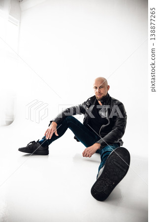 Man sits on floor wearing black leather jacket and jeans in a bright room with white walls Man sits on floor wearing black leather jacket and jeans in a bright room with white walls 138877265