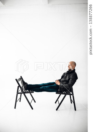 Man sits on a chair with legs stretched out on another chair in a simple studio space Man sits on a chair with legs stretched out on another chair in a simple studio space 138877266