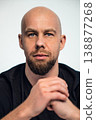 Person with shaved head sits with hands clasped in front of a plain background indoors 138877268