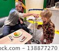 Man serves food to young girl at a table in a bright indoor space during evening hours 138877270