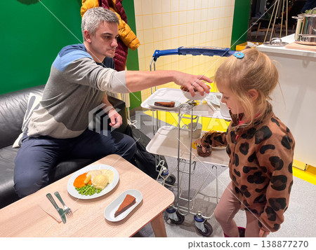 Man serves food to young girl at a table in a bright indoor space during evening hours 138877270