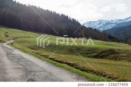 Countryside road leading through green meadow toward mountains 138877409