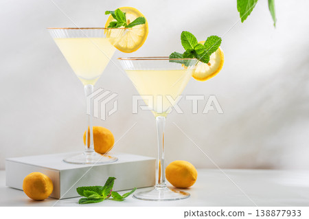 Bright and refreshing cocktail with two glasses of limoncello spritz, garnished with fresh lemon slices and mint sprigs on a white surface, with green foliage in the background Bright and refreshing cocktail with two glasses of limoncello spritz, garnished with fresh lemon slices and mint sprigs on a white surface, with green foliage in the background 138877933