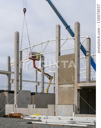 Crane lifting steel truss as worker installs frame on concrete columns at construction site 138878037