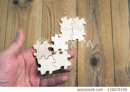 Human hand holding puzzle pieces trying to connect them on a wooden table. Human hand holding puzzle pieces trying to connect them on a wooden table. 138878109
