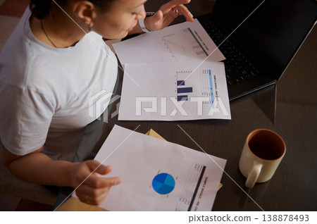 Woman Working From Home Reviewing Business Reports And Charts At Laptop With Coffee Woman Working From Home Reviewing Business Reports And Charts At Laptop With Coffee 138878493
