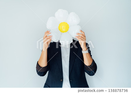 Business woman hides head behind large daisy flower balloon, symbolizing positivity and optimism in business. Concept of cheerful brand image and power of presenting simplicity, growth, fresh ideas. Business woman hides head behind large daisy flower balloon, symbolizing positivity and optimism in business. Concept of cheerful brand image and power of presenting simplicity, growth, fresh ideas. 138878736