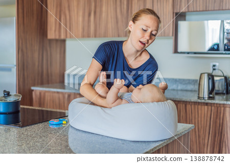 Mother talking to baby lying in cozy lounger on kitchen table while cooking at home. Caring mom interacting with infant during daily routine. Parenting, multitasking and family life concept Mother talking to baby lying in cozy lounger on kitchen table while cooking at home. Caring mom interacting with infant during daily routine. Parenting, multitasking and family life concept 138878742