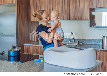Mother holding baby while cooking lunch in kitchen at home. Caring mom multitasking with infant during daily routine. Parenting, motherhood, healthy lifestyle and family life concept 138878745