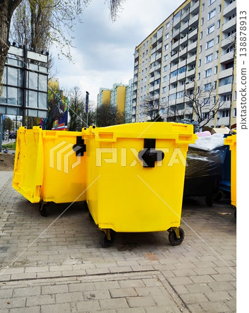 Bright yellow garbage bins are positioned on a paved area in an urban environment, a communal waste collection area 138878913