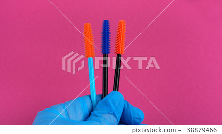 Hand in blue glove holding colorful mascara wands for eyelash extension on pink background Hand in blue glove holding colorful mascara wands for eyelash extension on pink background 138879466