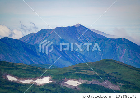A distant view of Hokkaido's high mountains, with its blue, layered ridgelines and snowfields. A spectacular landscape representative of Hokkaido's magnificent nature. A landscape photograph capturing the majestic natural beauty of Hokkaido. 138880175