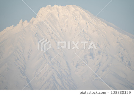 A close-up of Mount Rishiri with its glacial ridges and steep slopes. A spectacular view that represents the magnificent nature of Hokkaido. The snow-covered mountaintop shines against a clear blue sky. A close-up of Mount Rishiri with its glacial ridges and steep slopes. A spectacular view that represents the magnificent nature of Hokkaido. The snow-covered mountaintop shines against a clear blue sky. 138880339