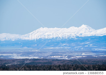 A distant mountain range of snow-capped peaks visible across the winter plains. A breathtaking view that represents the magnificent nature of Hokkaido. The snow-covered mountain peaks and the clear blue sky of winter. A distant mountain range of snow-capped peaks visible across the winter plains. A breathtaking view that represents the magnificent nature of Hokkaido. The snow-covered mountain peaks and the clear blue sky of winter. 138880353