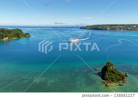 一艘渡輪航行在沖繩古利島周圍的海面上，船尾拖著長長的船尾波，從空中俯瞰碧綠的大海。 138881227