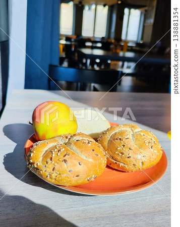 Two Kaiser rolls topped with sesame seeds rest on a red plate, accompanied by an apple at a cafe. Simple breakfast Two Kaiser rolls topped with sesame seeds rest on a red plate, accompanied by an apple at a cafe. Simple breakfast 138881354