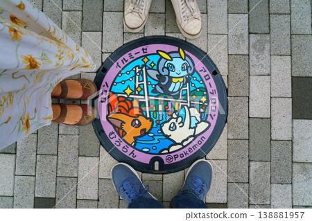 Muroran, Hokkaido, Japan - Oct 4 2024, A close-up view of a manhole with a color image of three cute Pokemon on background of Hakucho Bridge, with three people's feet standing around, Muroran, Japan Muroran, Hokkaido, Japan - Oct 4 2024, A close-up view of a manhole with a color image of three cute Pokemon on background of Hakucho Bridge, with three people's feet standing around, Muroran, Japan 138881957