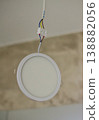 A modern round white LED recessed panel light hanging by exposed electrical wires and a terminal block from a ceiling during installation. High quality photo 138882056