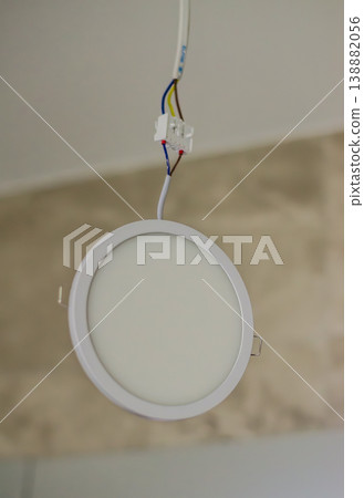 A modern round white LED recessed panel light hanging by exposed electrical wires and a terminal block from a ceiling during installation. High quality photo A modern round white LED recessed panel light hanging by exposed electrical wires and a terminal block from a ceiling during installation. High quality photo 138882056