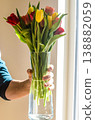 A person holding a vase of vibrant, colorful spring tulips illuminated by bright natural sunlight next to a window. Floral gift concept. . High quality photo 138882059