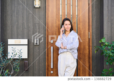 A Japanese woman smiles as she talks on her smartphone in front of the entrance of a detached house before going out. 138882729