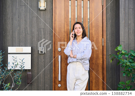 A Japanese woman smiles as she talks on her smartphone in front of the entrance of a detached house before going out. A Japanese woman smiles as she talks on her smartphone in front of the entrance of a detached house before going out. 138882734