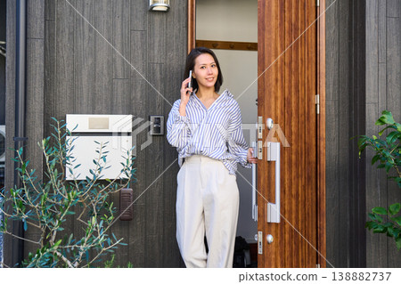 A Japanese woman opens the front door of a house and goes out while talking on her smartphone. A Japanese woman opens the front door of a house and goes out while talking on her smartphone. 138882737