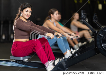Positive Latina girl training with rowing machine in sports hall Positive Latina girl training with rowing machine in sports hall 138884429