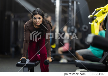 Latina woman doing dumbbell rows near bench in hall 138884463