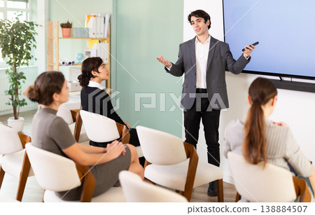 Portrait of young businessman standing near interactive board and showing presentation with remote control for group of students sitting in chairs in auditorium Portrait of young businessman standing near interactive board and showing presentation with remote control for group of students sitting in chairs in auditorium 138884507