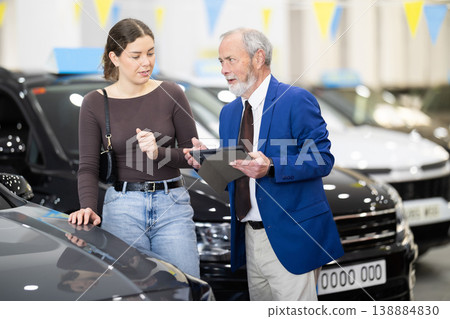 Elderly male car seller in suit walking around car salon with woman who wants to buy car and talking about specifications of cars Elderly male car seller in suit walking around car salon with woman who wants to buy car and talking about specifications of cars 138884830