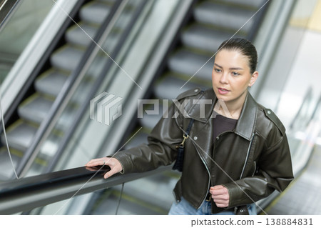 Positive young woman in black leather jacket riding up an escalator at metro station 138884831