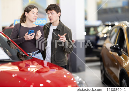 Young couple is standing in a car dealership and looking at the car with gadget in hands Young couple is standing in a car dealership and looking at the car with gadget in hands 138884941
