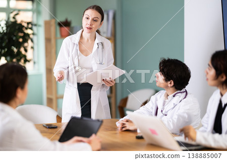Confident young woman doctor standing near table where sitting medical interns talking and discussing ideas in medical office Confident young woman doctor standing near table where sitting medical interns talking and discussing ideas in medical office 138885007
