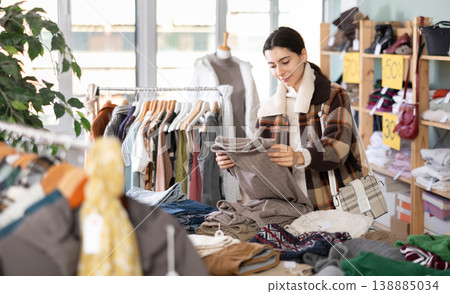 Focused young girl client selecting trousers in clothing shop. Winter clothing collection Focused young girl client selecting trousers in clothing shop. Winter clothing collection 138885034