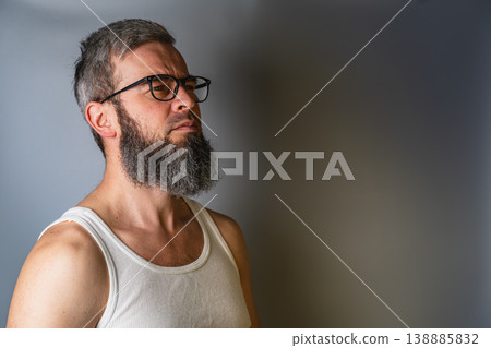 Man with beard stands against a gray wall wearing a tank top Man with beard stands against a gray wall wearing a tank top 138885832