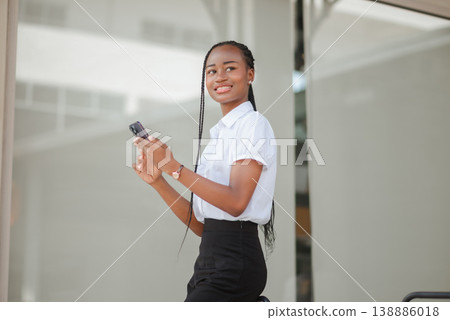 Young African professional woman standing outdoors and holding smartphone while looking away, concept of mobile communication, confidence, modern business lifestyle and remote work flexibility. 138886018