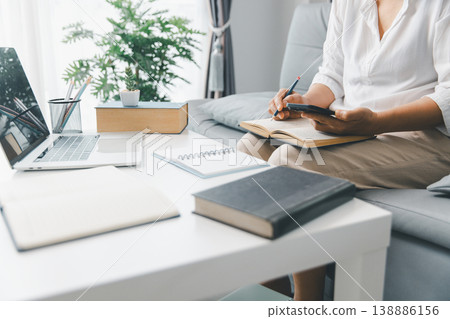 Home study scene: person on sofa reading open book while checking smartphone with laptop and notebooks on white table. Education, online learning, homework and exam prep concept. student, education 138886156