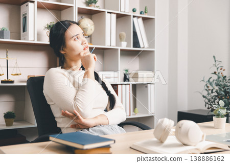Young Asian woman thinking at home office desk with pen, books and headphones, studying or planning ideas. Thoughtful student, problem solving, online learning and productivity concept. 138886162