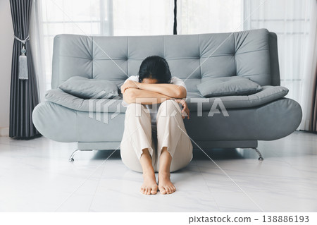 Depressed woman sitting on floor in front of gray sofa, head down on arms and knees, barefoot at home in bright living room. Mental health anxiety, sadness, burnout, loneliness concept. depression Depressed woman sitting on floor in front of gray sofa, head down on arms and knees, barefoot at home in bright living room. Mental health anxiety, sadness, burnout, loneliness concept. depression 138886193