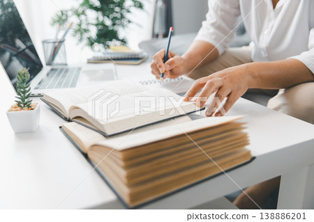 Hands studying at home, reading open textbooks while taking notes beside laptop on bright white desk with stationery and plant. Education, online learning, homework and exam prep concept. learning Hands studying at home, reading open textbooks while taking notes beside laptop on bright white desk with stationery and plant. Education, online learning, homework and exam prep concept. learning 138886201