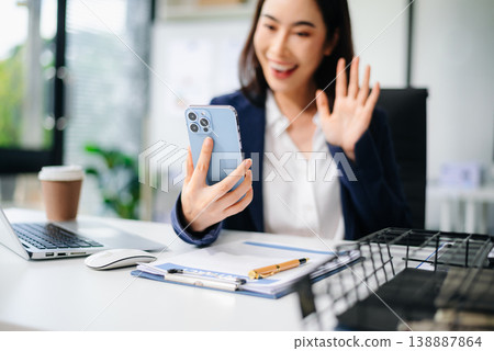 Asian businesswoman waving and smiling on smartphone video call from office desk with laptop and coffee. Asian businesswoman waving and smiling on smartphone video call from office desk with laptop and coffee. 138887864