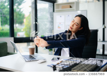 Asian businesswoman stretching arms in office with tired expression, relieving muscle tension from long work hours. 138887969