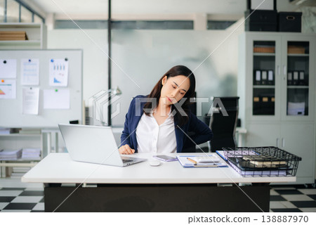 Asian businesswoman stretching arms in office with tired expression, relieving muscle tension from long work hours. 138887970