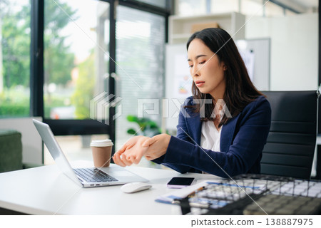 Asian businesswoman stretching arms in office with tired expression, relieving muscle tension from long work hours. 138887975