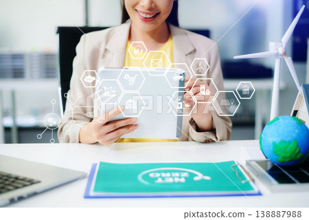 Asian woman using tablet with futuristic eco interface showing CO2, battery, wind, and recycle icons. Concept of smart sustainability, net zero tech, and green innovation. 138887988
