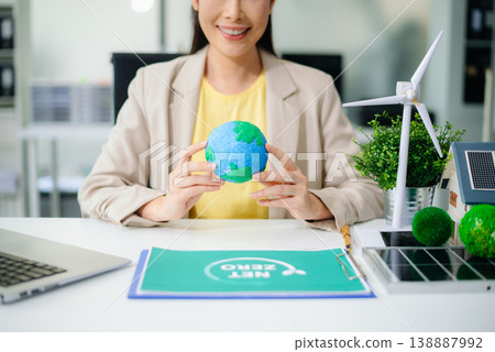 Asian woman in office holding earth model beside solar panels and wind turbine. Concept of sustainability, climate change, and clean energy innovation. Asian woman in office holding earth model beside solar panels and wind turbine. Concept of sustainability, climate change, and clean energy innovation. 138887992