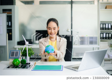 Asian woman in office holding earth model beside solar panels and wind turbine. Concept of sustainability, climate change, and clean energy innovation. Asian woman in office holding earth model beside solar panels and wind turbine. Concept of sustainability, climate change, and clean energy innovation. 138888009